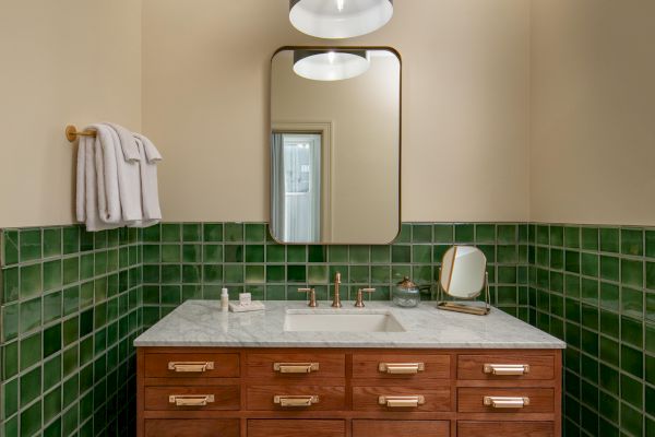 A bathroom with green tiles, a wooden vanity, a round mirror, and a black pendant light. Towels are hung on the wall to the left.
