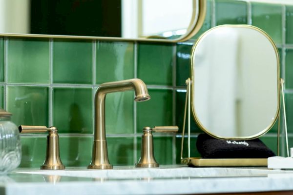 A bathroom counter with a gold faucet, round mirror, tissue box, glass jar, and toiletries set against green tiled walls.