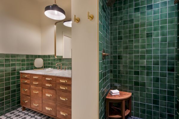 A bathroom with green tiled walls, a wooden vanity with brass handles, a sink, a round mirror, and a wooden stool with towels in the shower area.