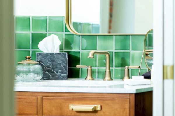 A bathroom sink with gold fixtures, green tiles, a marble tissue box, a glass jar, a mirror, and a soap tray is partially visible.