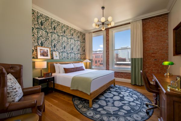 A stylish bedroom with a bed, patterned wallpaper, brick accent wall, desk, leather chair, and large windows with a city view, and a decorative rug.