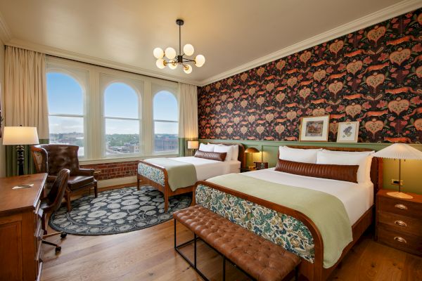 A hotel room features two beds, an ornate wallpaper, a desk, a leather chair, and large windows with a scenic view, along with a stylish chandelier.