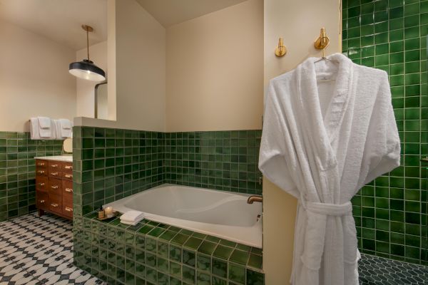 A bathroom with green tile, bathtub, white robe, towel rack, wooden drawers, and a hanging light fixture. Black and white patterned floor.