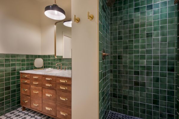 The image shows a bathroom with green tiled walls, a wooden vanity with brass handles, a sink, round mirror, pendant light, and green-tiled shower area.