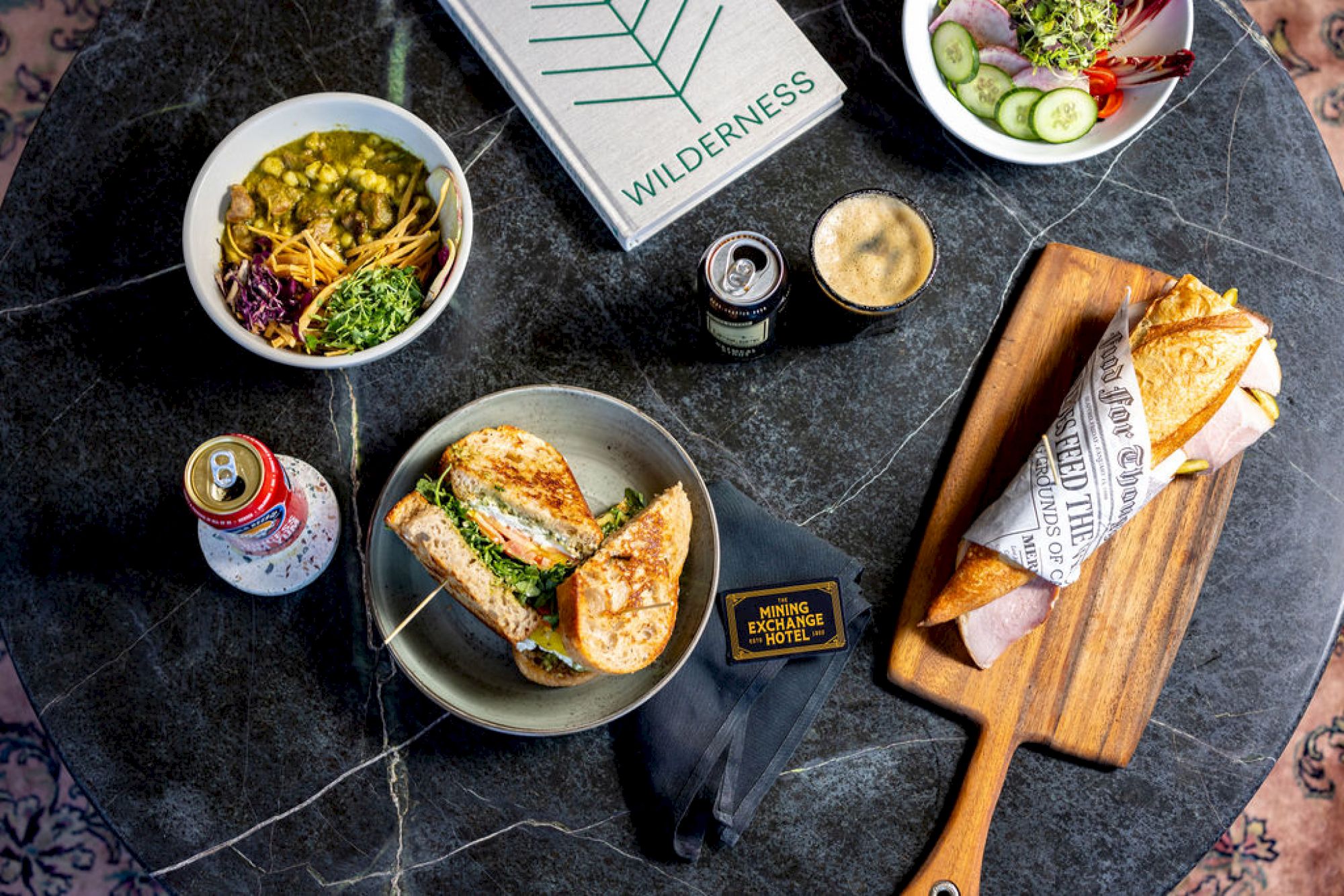 The image shows various food and drinks on a dark table, including sandwiches, a bowl, a can of drink, a coffee, a book, and a wooden board.