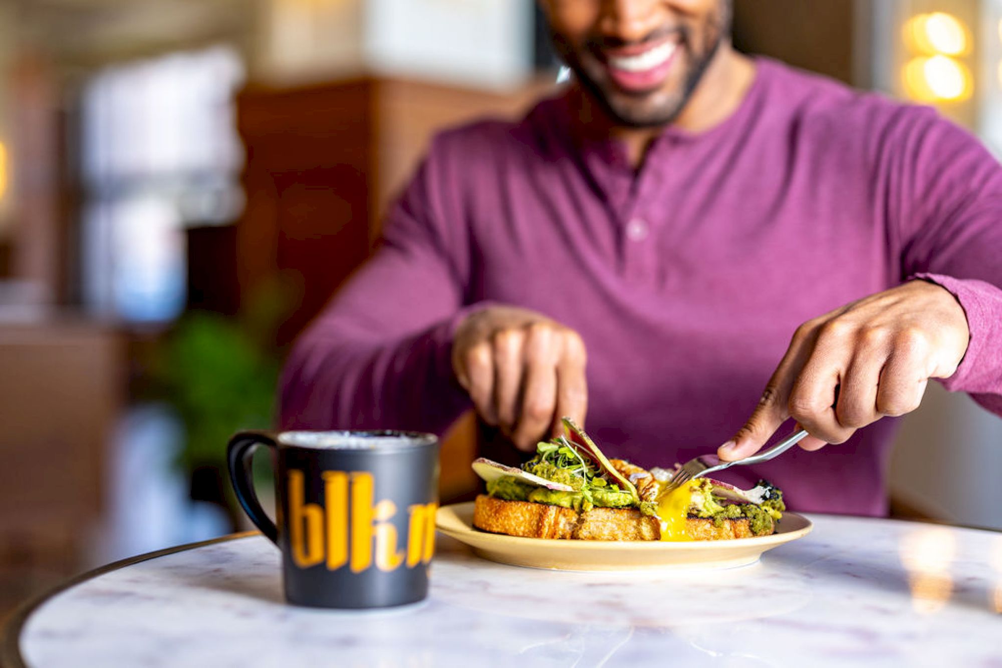 A person is cutting food on a plate while smiling, with a black mug that appears to say 