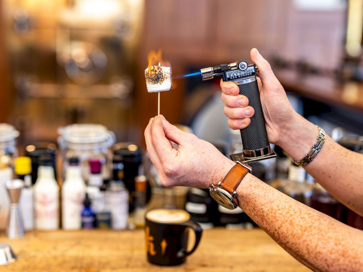 A person is using a torch to toast a marshmallow over a bar counter, with various bottles and a cup in the background.
