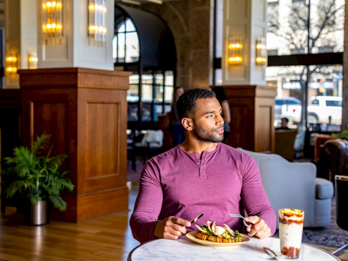 A man in a purple shirt sits at a table in a stylish restaurant, enjoying a meal and a dessert while looking off to the side.