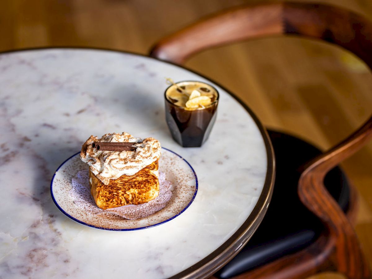 A marble table with a pastry topped with cream and chocolate, and a cup of coffee, beside a wooden chair.