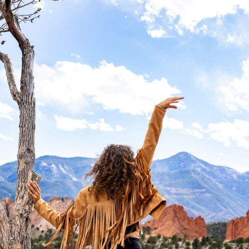 A person in a fringed jacket stands near a tree, raising their arm, with a scenic backdrop of mountains and a partly cloudy sky.
