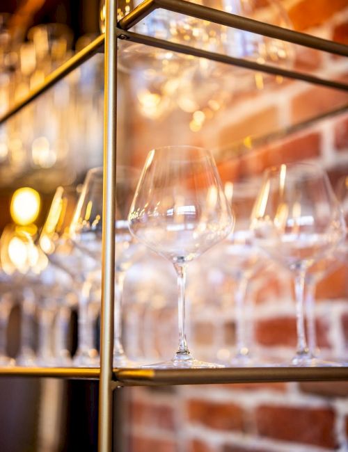 The image shows a collection of empty wine glasses neatly arranged on glass shelves against a brick wall backdrop.