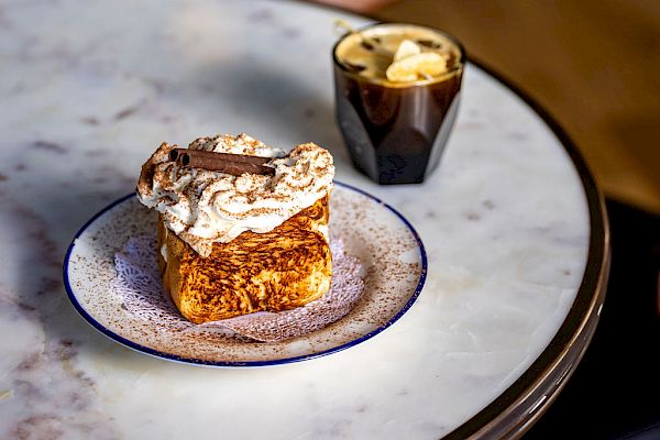 A marble table holds a piece of cake on a plate and a cup of coffee topped with cream.
