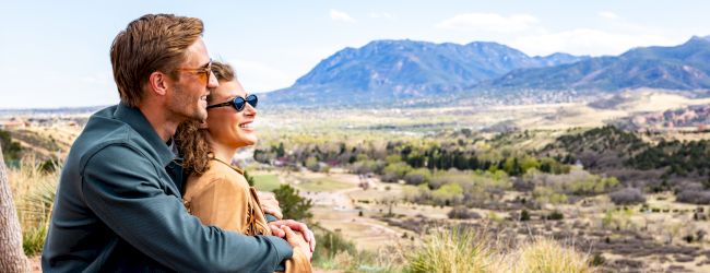 A couple sits outdoors, embracing while enjoying a scenic view of mountains and a valley on a sunny day, both wearing sunglasses.