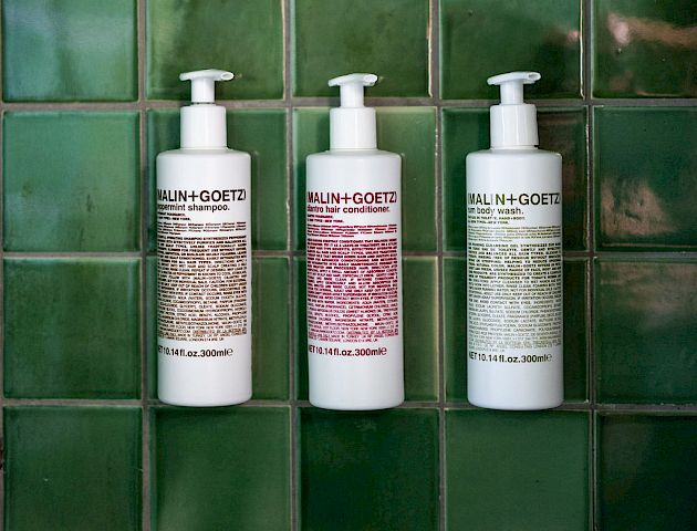 Three white pump bottles with text are mounted on a dark green tiled wall. They appear to be toiletries, likely shampoo, conditioner, and body wash.