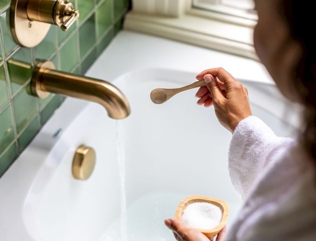 A person in a white robe is adding bath salts from a wooden spoon into a filling bathtub with a gold faucet in a bathroom with green tiles.