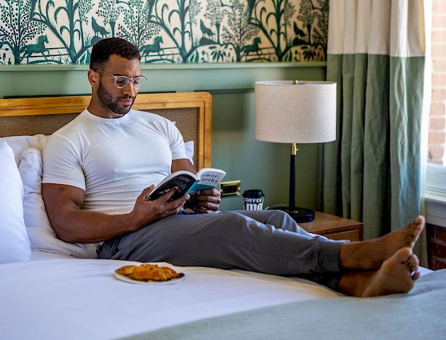A man is sitting on a bed, reading a book, with a snack next to him. There's a lamp on the bedside table and patterned wallpaper in the background.