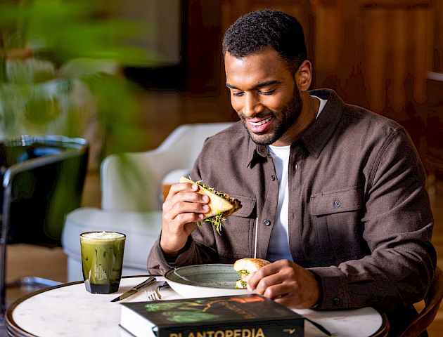 A man sits at a table with a smoothie and a sandwich, reading a book titled 