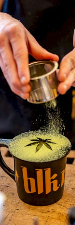 A person dusting green powder, likely matcha, over a mug with a leaf design on top. The mug has the letters 