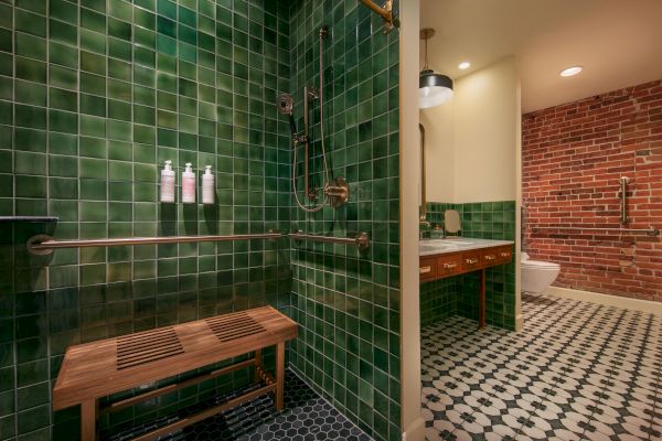 This image displays a stylish bathroom with green tiled walls, a wooden bench, and shower. It features a sink, brick wall, and patterned floor tiles.