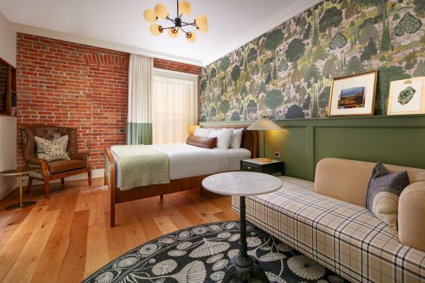 A cozy bedroom features a bed, a green sofa, a chair, a round table, patterned wallpaper, brick wall, framed art, and wooden flooring.