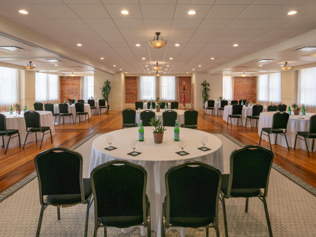 The image shows a conference or banquet hall with round tables and black chairs arranged in a spacious, well-lit room. There are small plants on the tables.