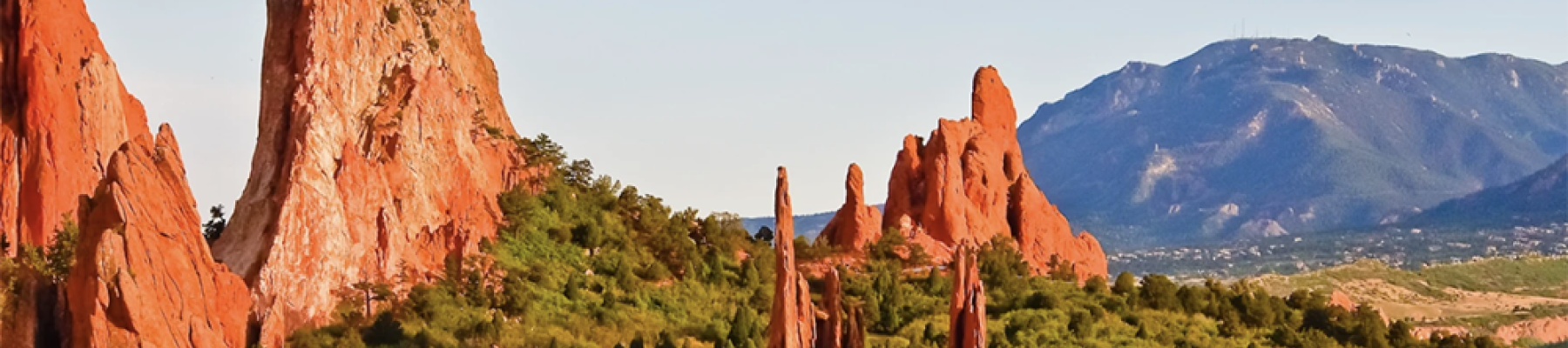 The image shows a landscape with towering red rock formations surrounded by lush greenery, with a mountain range in the background and a bright sky.