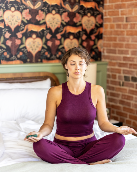 A person is meditating on a bed, dressed in a maroon outfit with a floral wallpaper and brick wall in the background.