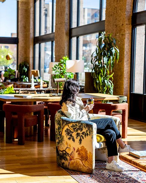 A woman is sitting in a cozy, brightly lit room with large windows, surrounded by plants and furniture, enjoying a beverage and looking outside.