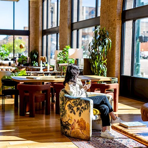 A woman is sitting in a cozy, brightly lit room with large windows, surrounded by plants and furniture, enjoying a beverage and looking outside.