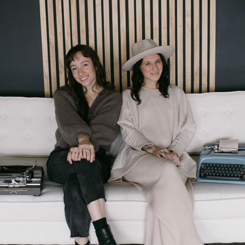 Two women are sitting on a white couch with typewriters on either side. The background features wooden slats and a dark wall.