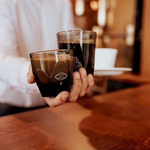 A person holding two glasses of dark coffee over a wooden surface, with a white shirt sleeve visible, in a cozy indoor setting.