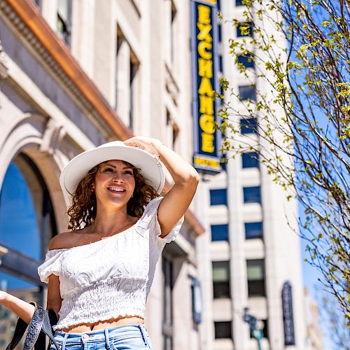 The image shows a woman in a white off-shoulder top and hat, smiling and holding her hat. She stands near a building with an 