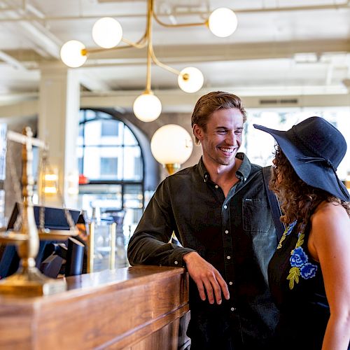 A man and woman are smiling and conversing in a warmly lit, elegant interior with wooden counters and modern light fixtures.