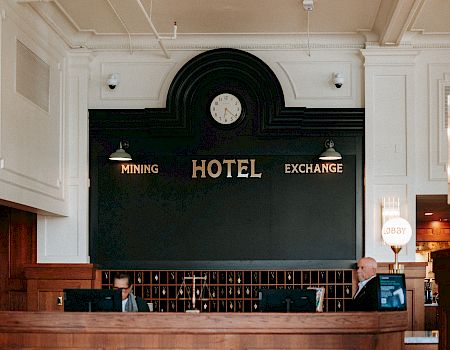 A hotel reception area with a wooden front desk, a clock above, and signs reading 