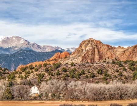 A scenic landscape featuring rugged mountainous terrain, a mix of red rock formations, and a distant snow-capped peak under a partly cloudy sky.